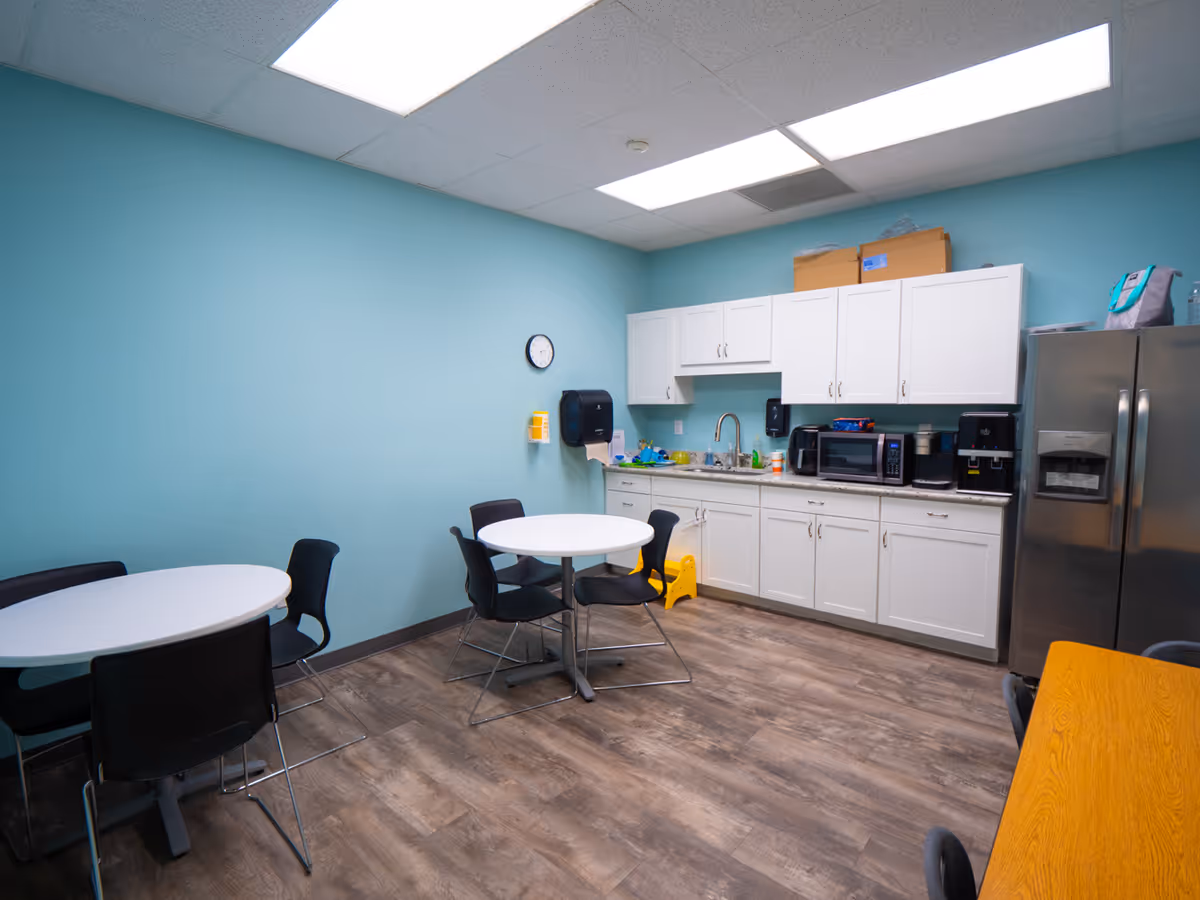 Teal-walled kitchenette with white cabinets, tables, chairs, and stainless steel appliances.
