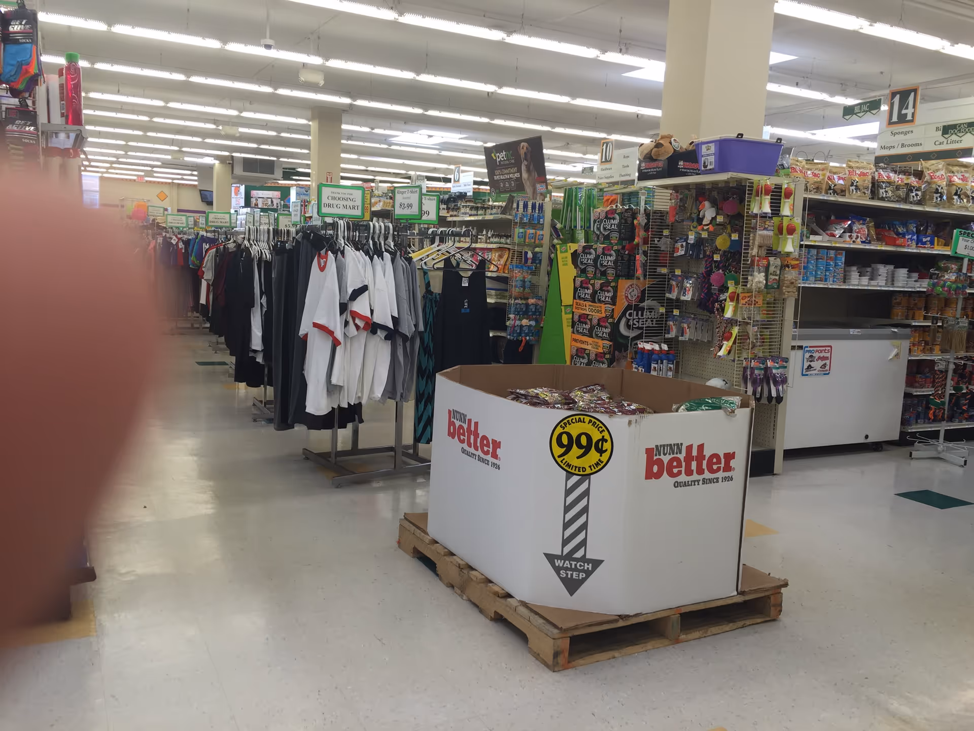 Inside a Discount Drug Mart store, clothing racks and merchandise surround a central display.