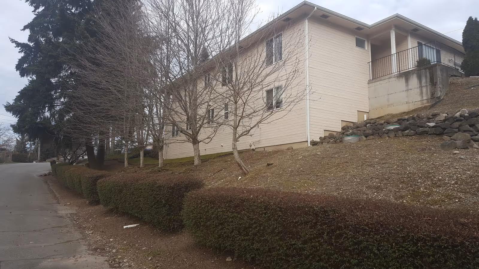 Exterior view of a beige building on a hillside with leafless trees and hedges.
