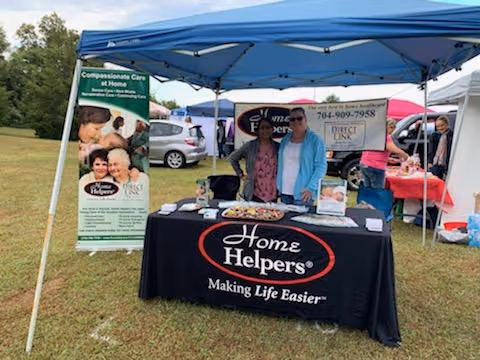 Two people stand behind a Home Helpers booth under a blue canopy at an outdoor event.