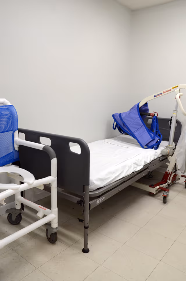 A clinical hospital bed with rails and a blue patient chair in a small, empty room.