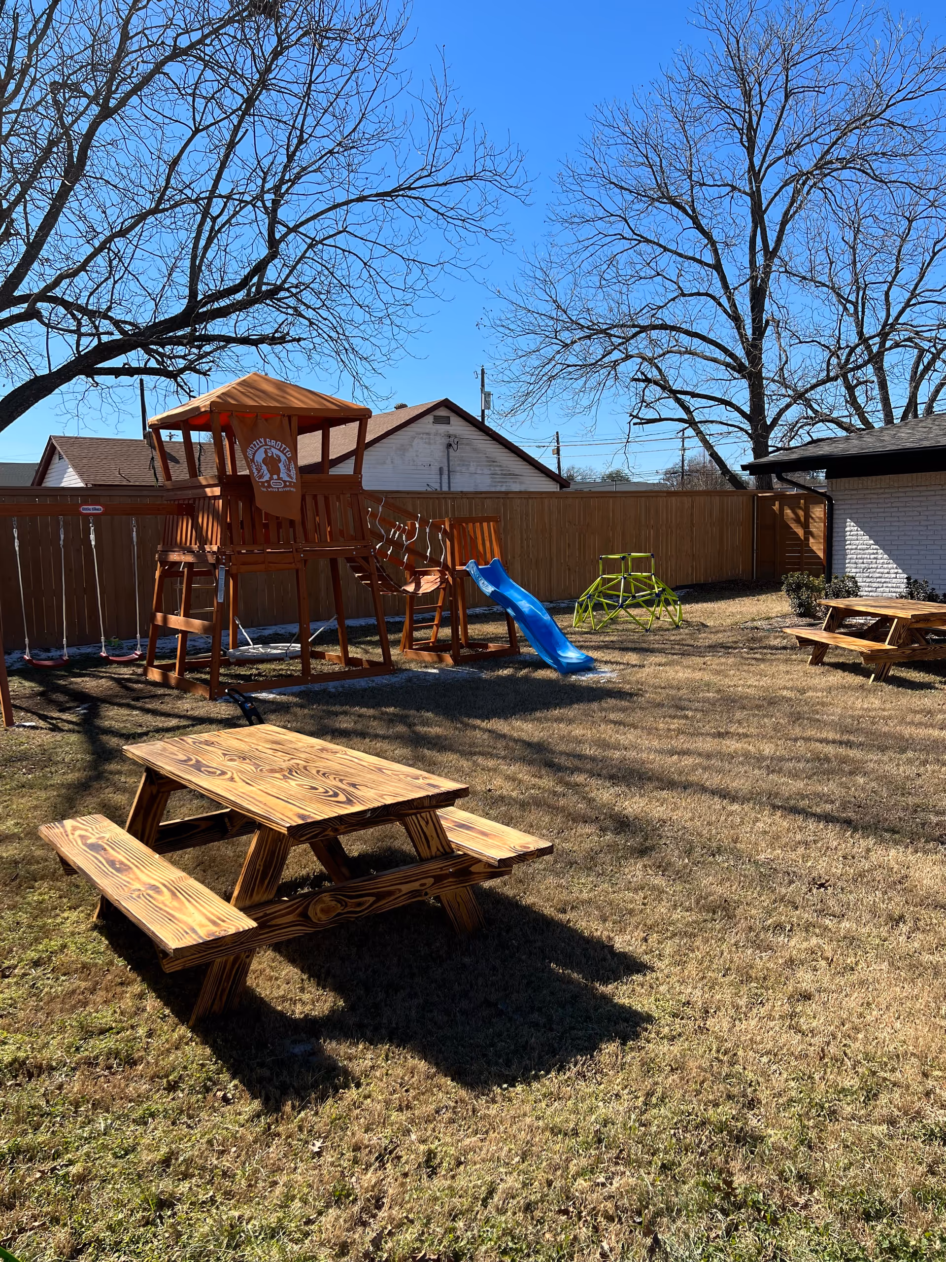 Backyard playground with a wooden playset, blue slide, swings, picnic table, and bare trees.