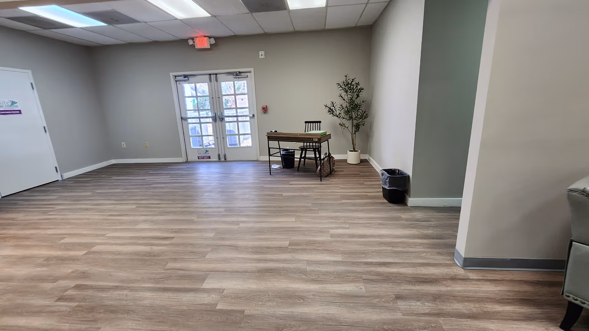 An empty interior room with a doorway, small desk, chair, potted plant, and trash bin.