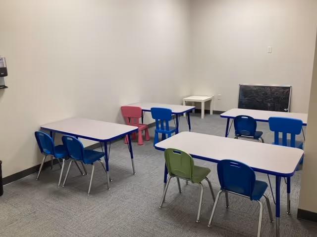 Activity room with multiple small tables and colorful chairs against neutral walls.