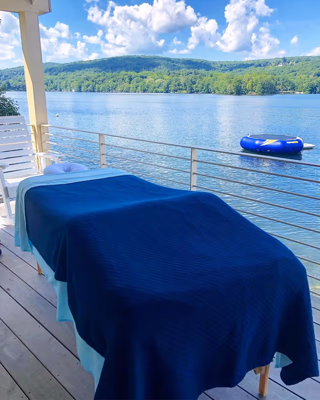 Outdoor massage table on a lakeside deck with blue blanket, overlooking calm water.