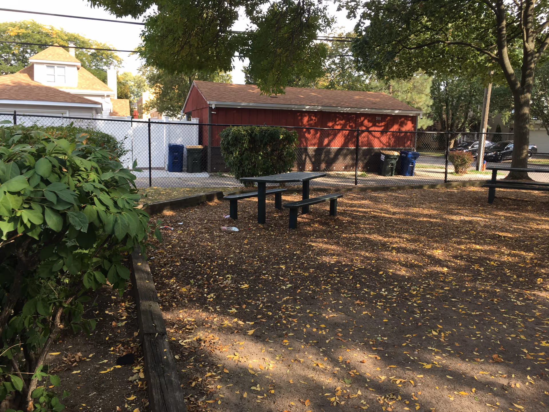 Outdoor communal area with picnic tables, benches, a chain-link fence, red shed, and trees.