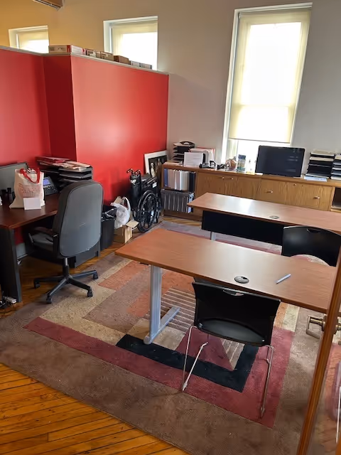 Interior office area with desks, chairs, computer, a wheelchair, and a red storage cabinet.