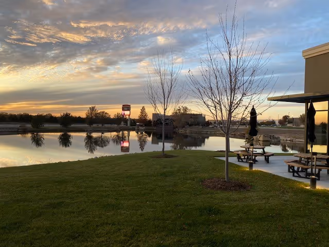 Sunset over a grassy lawn and pond with outdoor tables and bare trees beside a building.