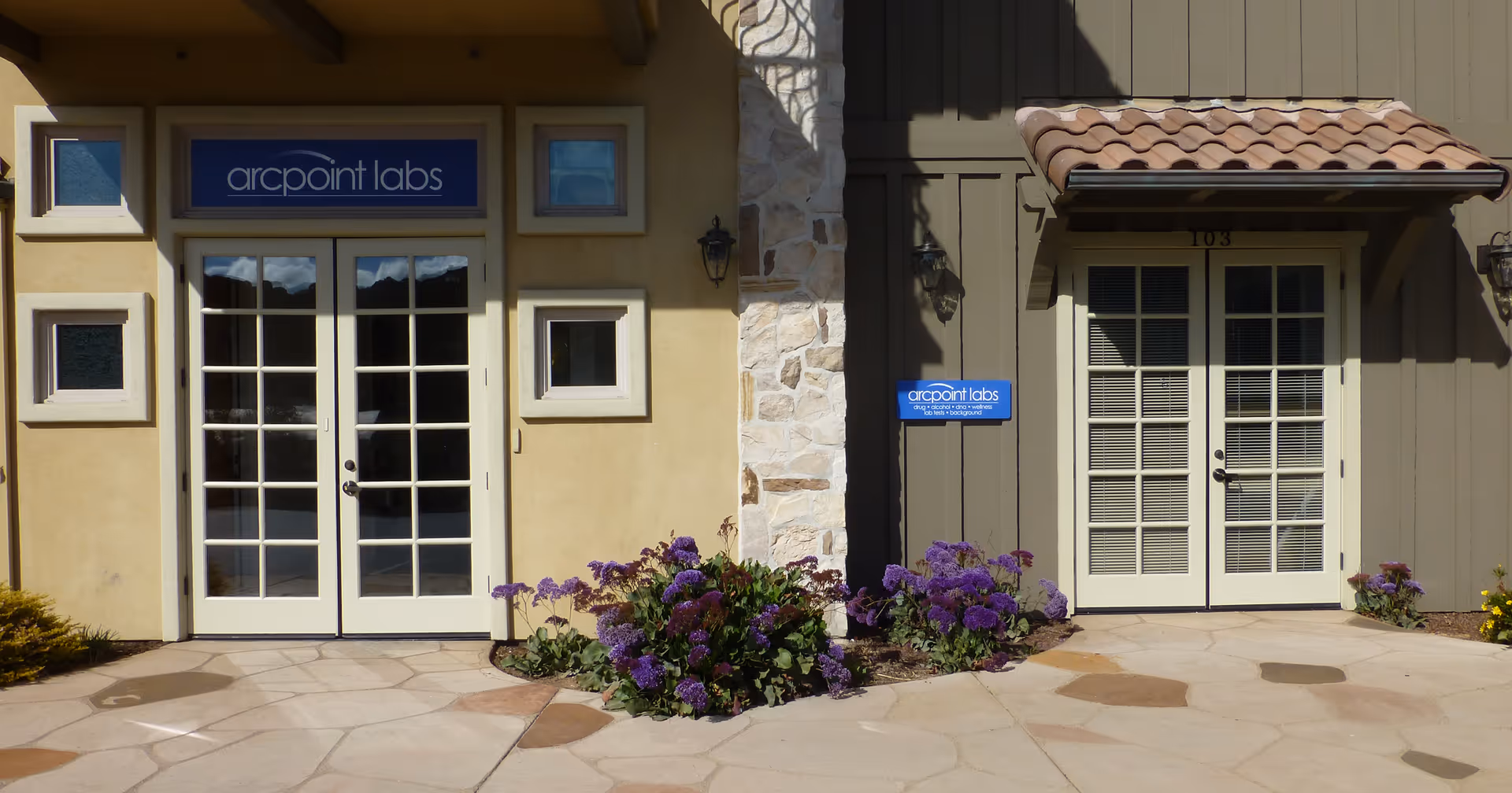 Two glass doors flank a stone pillar, with purple flowers in front.