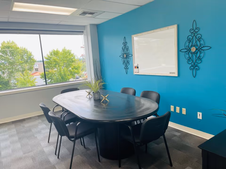 A teal-walled conference room with a round table and black chairs by a large window.