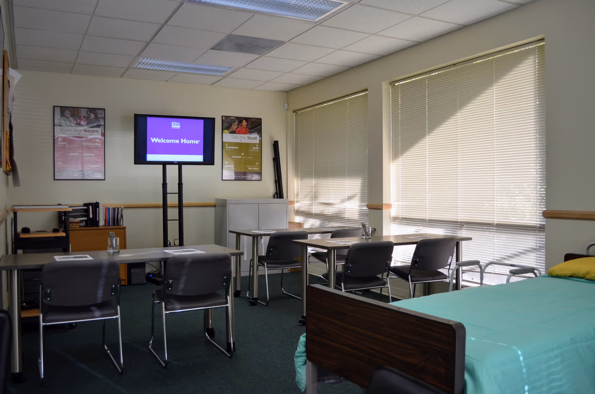 A small conference room in a senior facility with tables, chairs, a TV, and a bed.