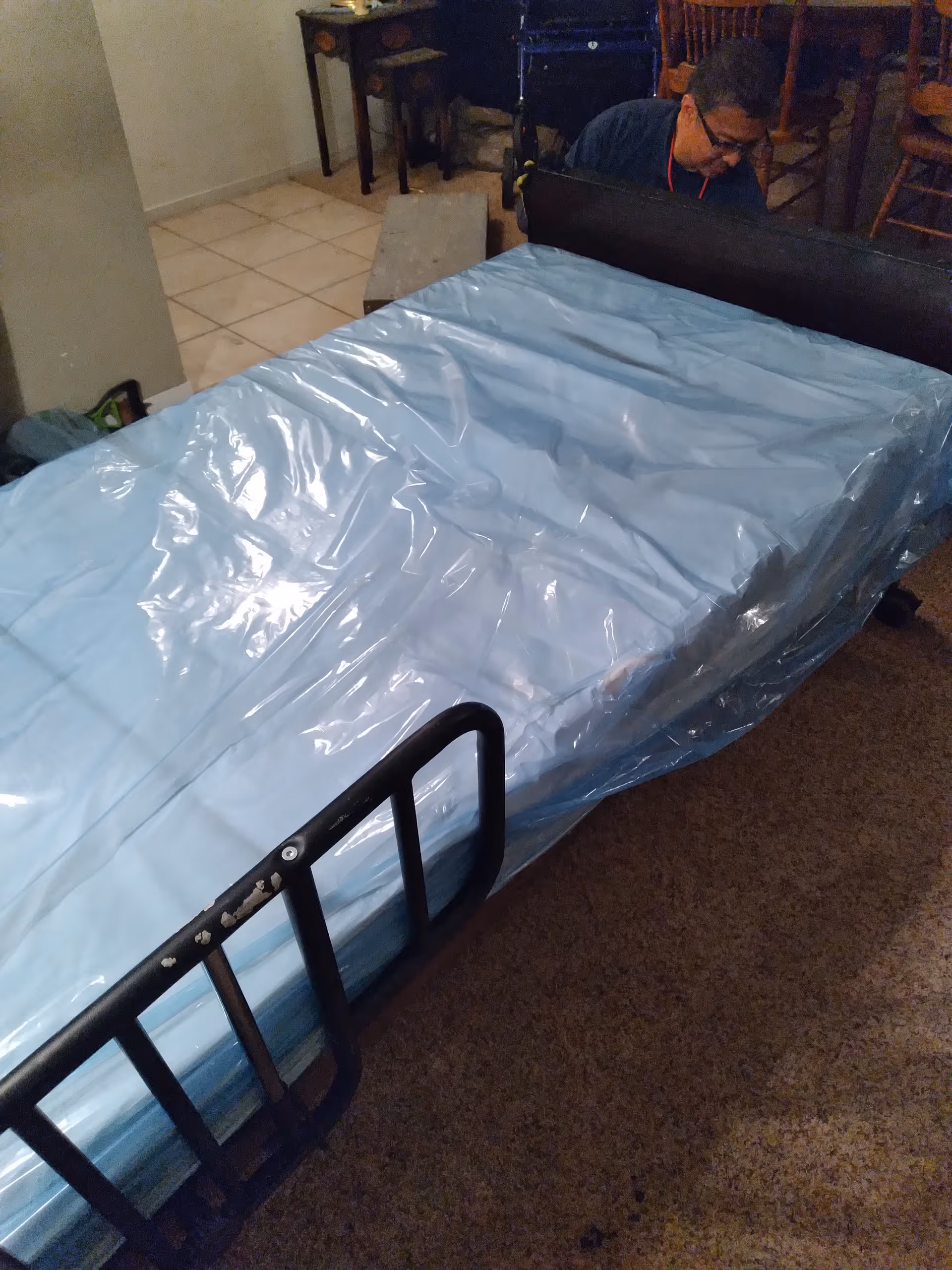 Hospital-style bed with blue plastic cover in a care room, a person at the far end.