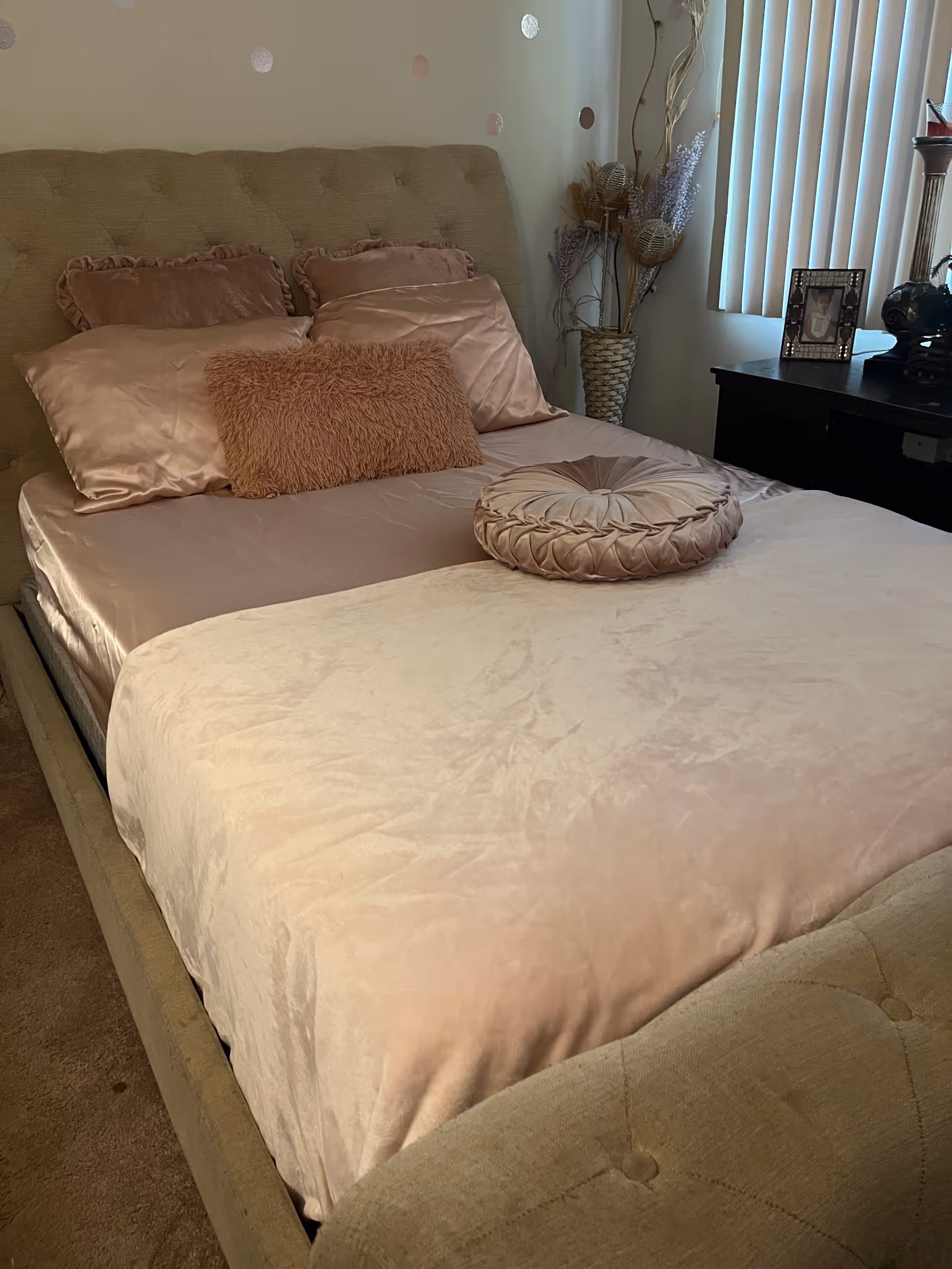 Soft pink bedding with tufted headboard, decorative pillows, and a nightstand beside a window.