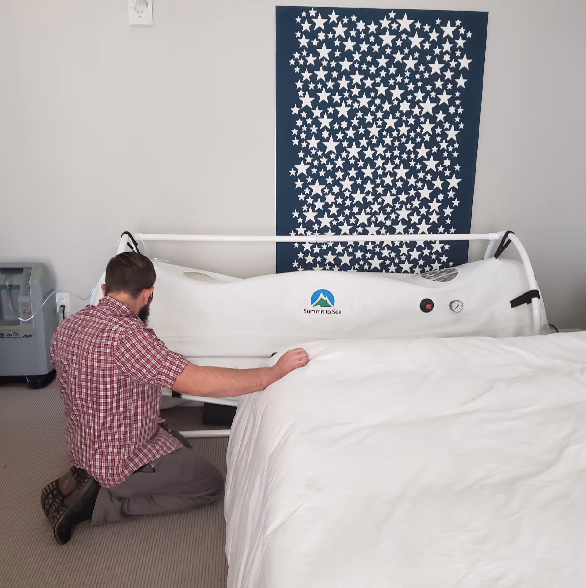 Man kneels beside a white medical bed in a bright room with star-pattern wall art.