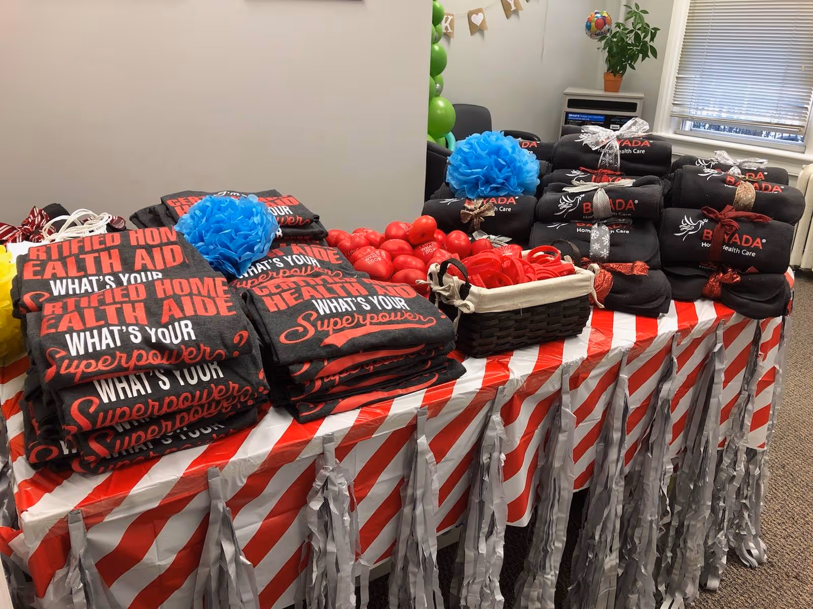 Table covered with red-striped cloth displays BAYADA-branded blankets and gifts in a room.