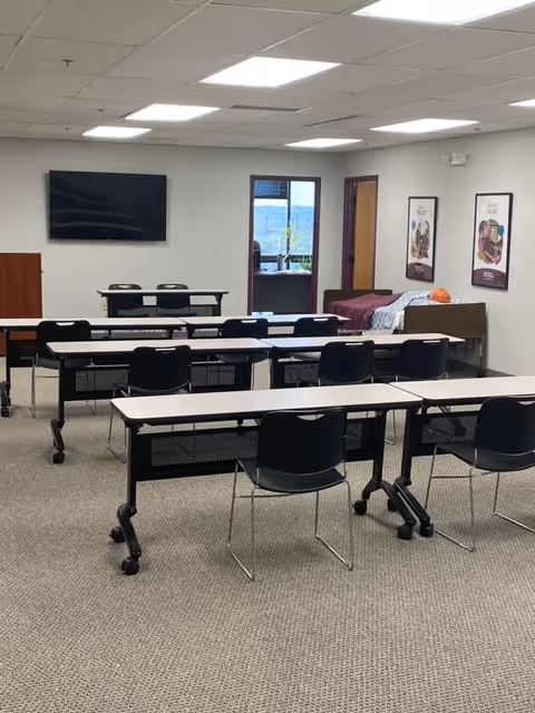 Classroom-style interior with rows of desks and chairs, a TV, and a bed in the back.