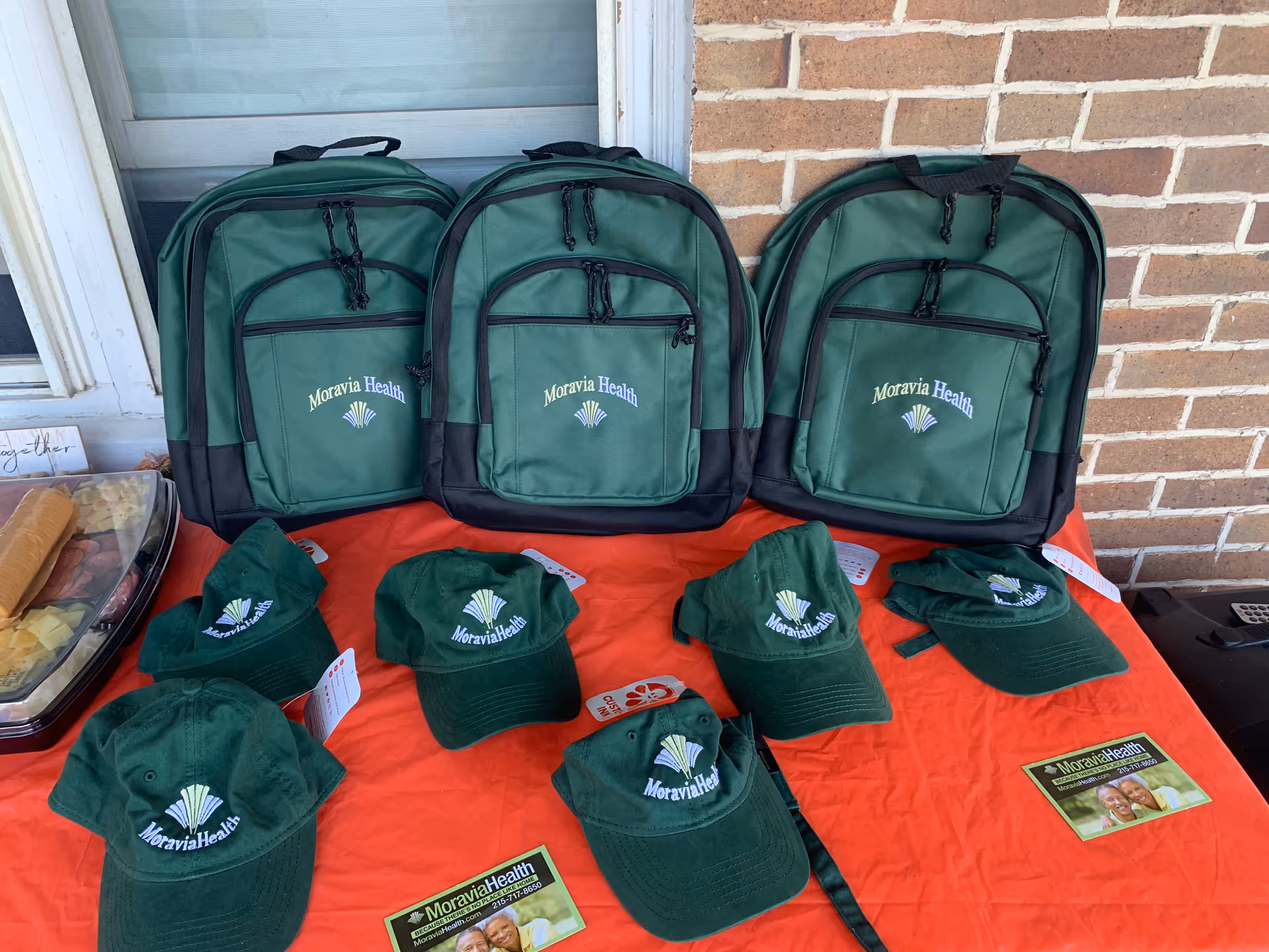 Outdoor display of Moravia Health backpacks and caps on a red tablecloth by brick wall.