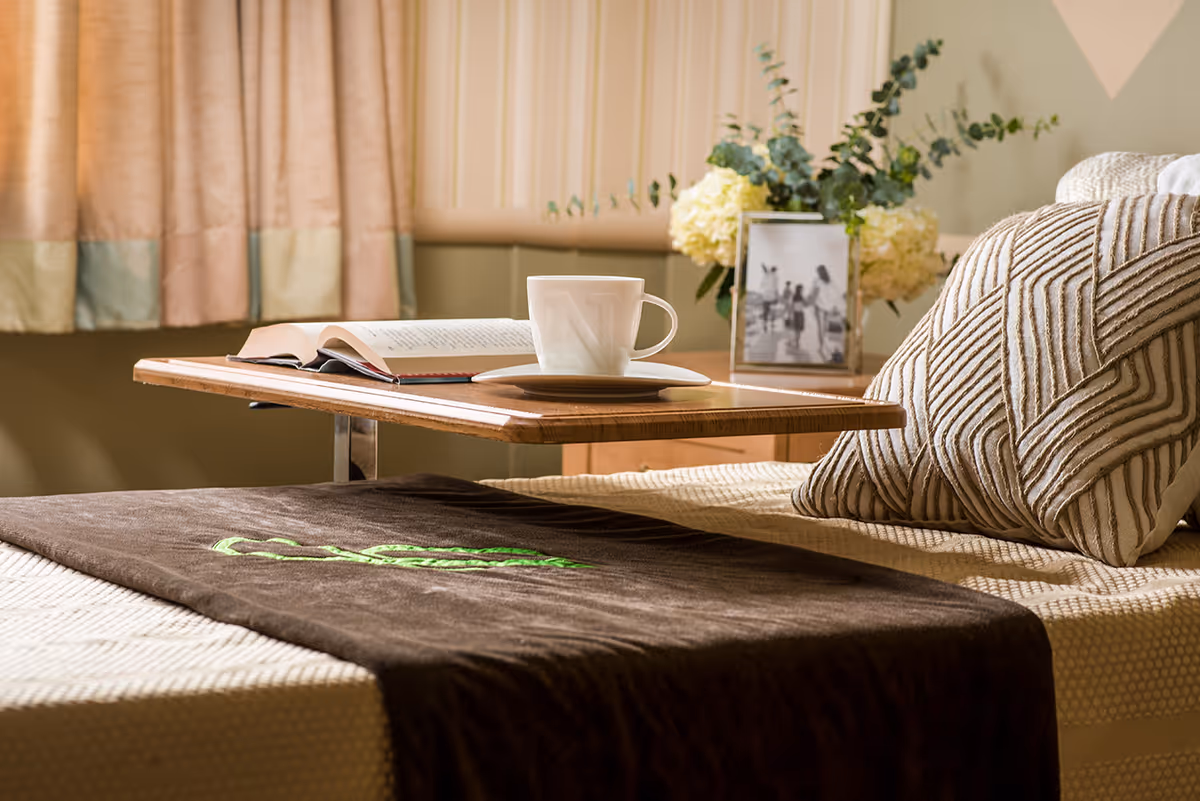 Bedside area of a senior living room with a tray table, mug, and open book.