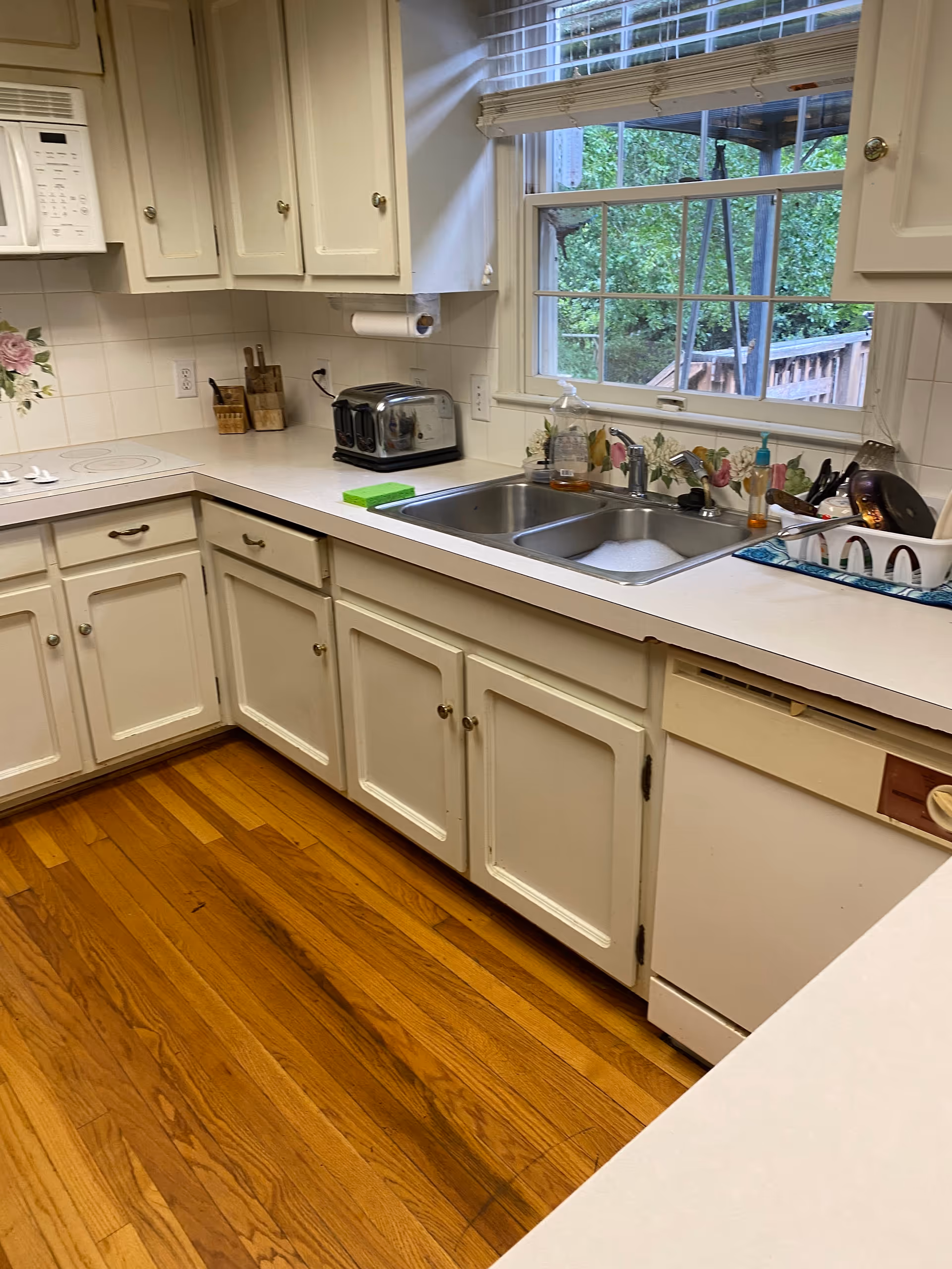 Bright white kitchen with cabinets, sink, toaster, and a window overlooking greenery.