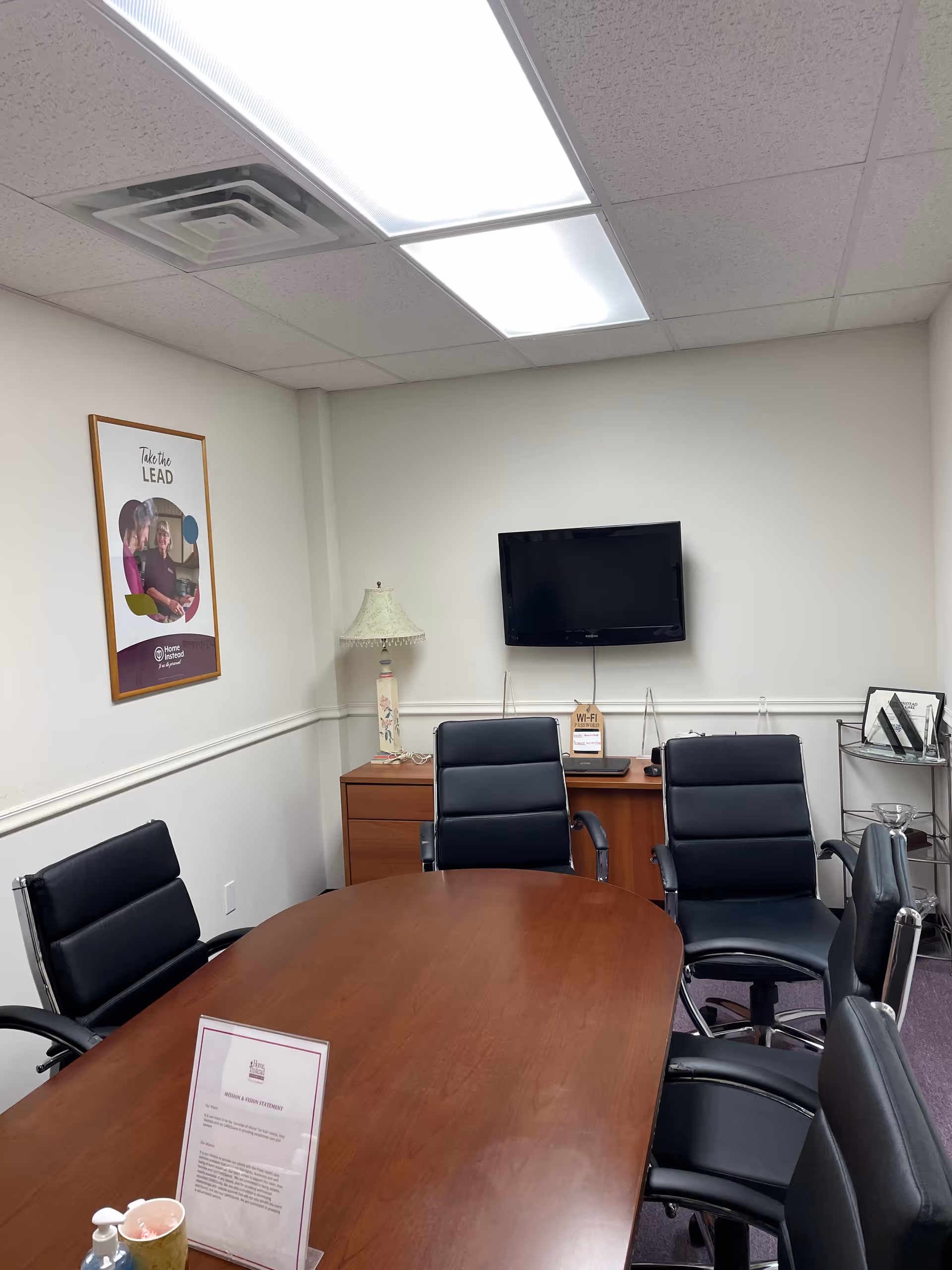 A conference room with a wooden oval table, black chairs, and a wall-mounted TV.