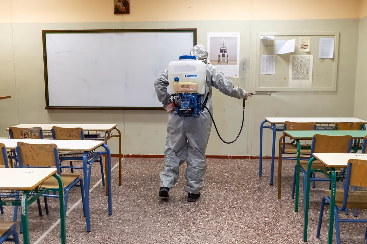 A person in protective suit disinfects a classroom with a backpack sprayer among desks.