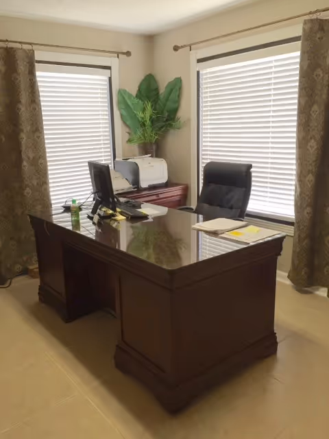 A dark wooden office desk with chair, printer, plant, and large window blinds.