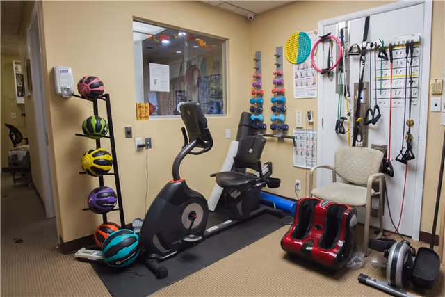 Small indoor rehabilitation gym with exercise bike, treadmill, weights, and therapy chair.
