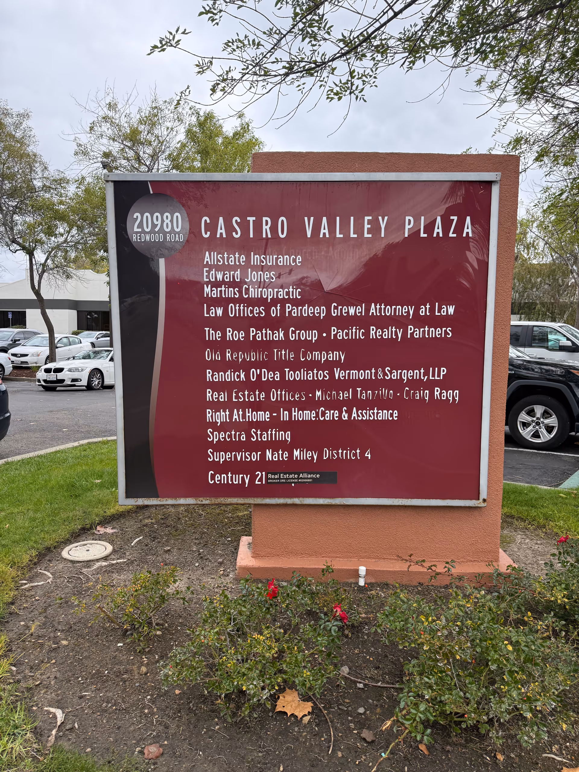 Large maroon plaza sign listing tenants, with cars and trees in background.