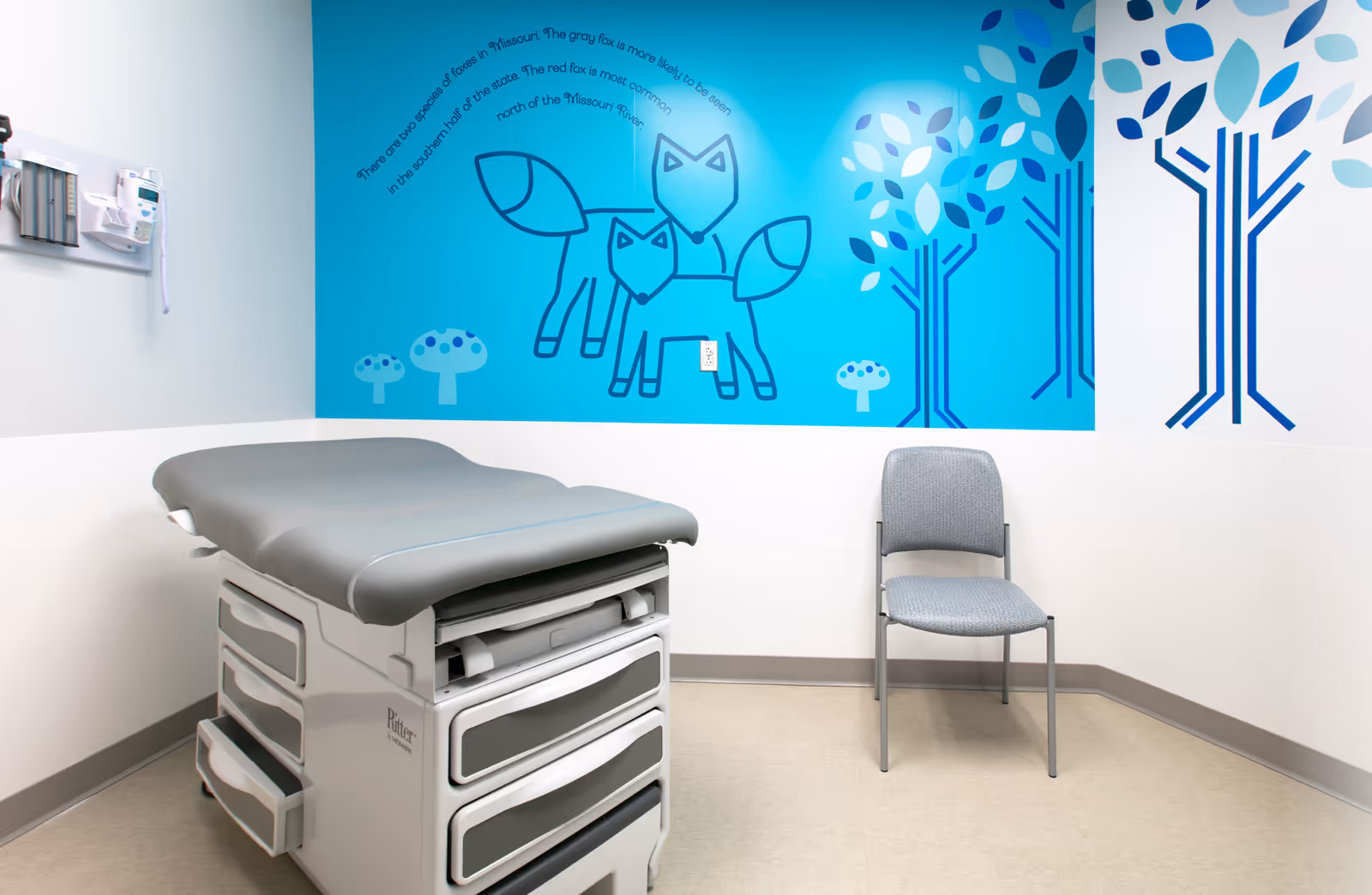 A medical examination table and chair in a bright clinic room with blue mural.