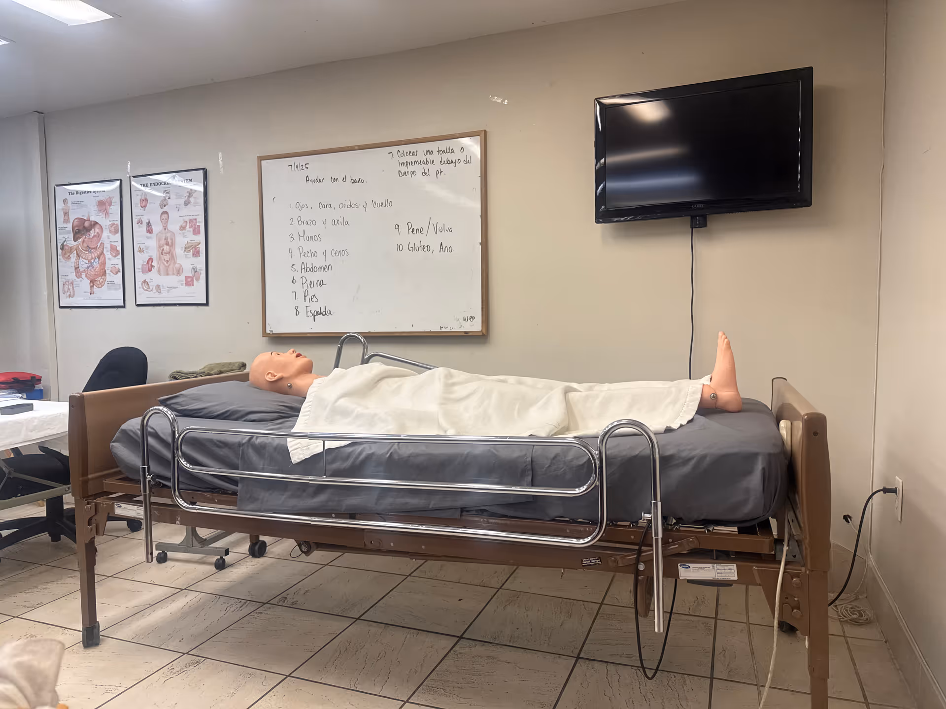 Hospital-style bed with mannequin under a sheet in a clinical room with a whiteboard and TV.
