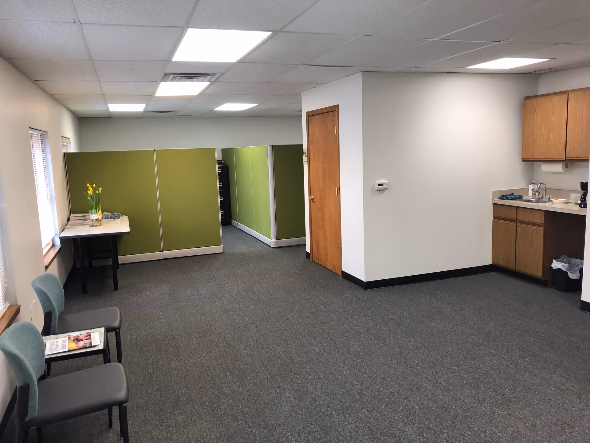 Interior common area with green cubicle partitions, a table with flowers, and a kitchenette.
