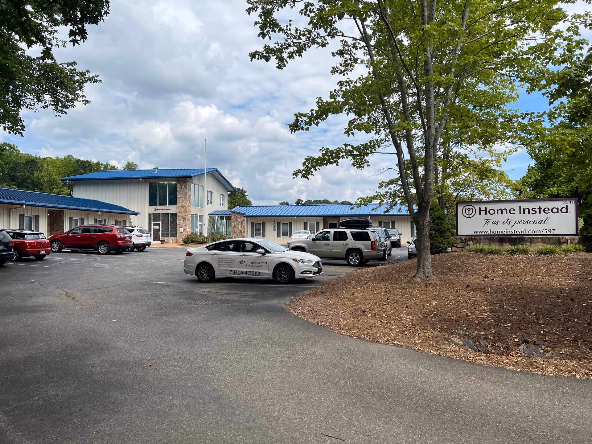Exterior view of a Home Instead facility with parked cars and a sign.