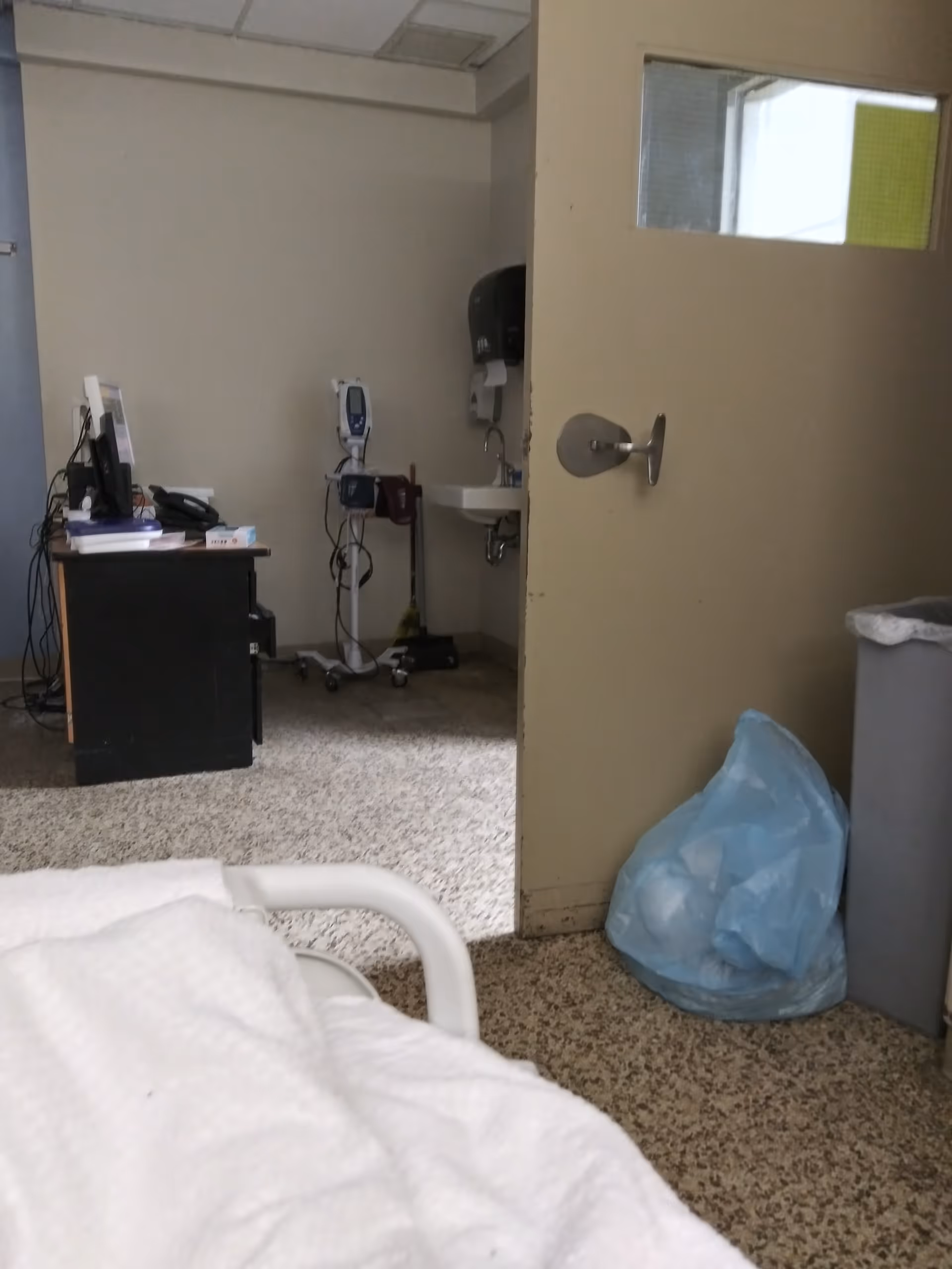 Hospital room interior showing desk with computer, medical devices, sink, and bed rail.