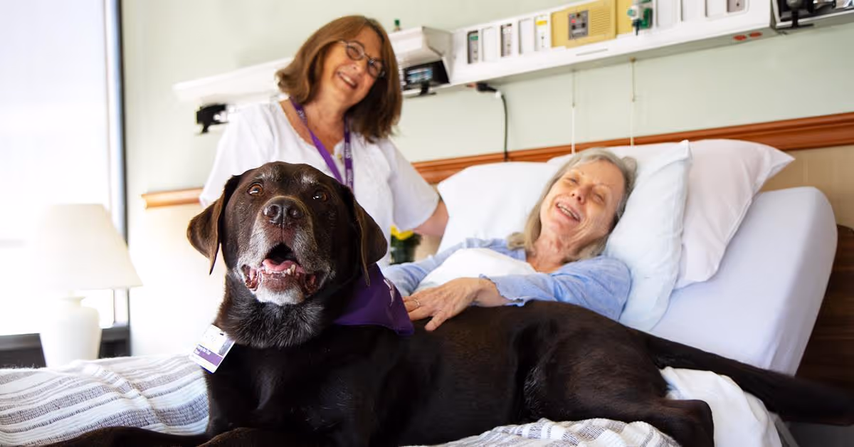 Smiling nurse beside a patient in bed with a large black dog in a hospital room.