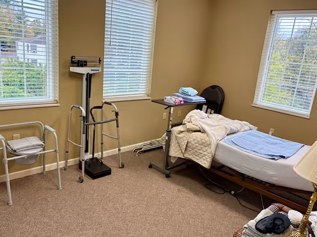 Care room with a bed, walker, scale, and overbed table by windows.