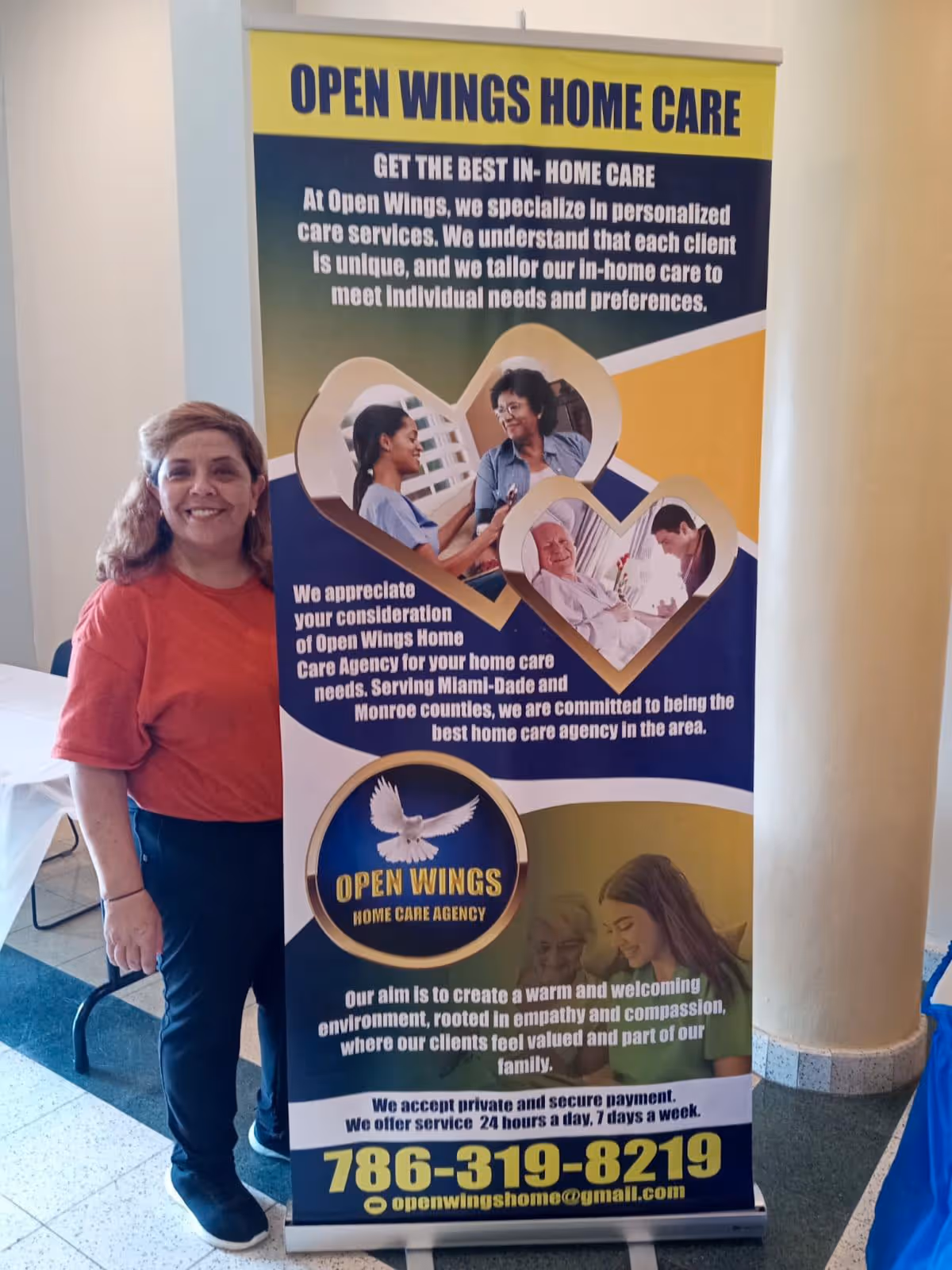 A smiling woman stands beside a large Open Wings Home Care banner indoors.
