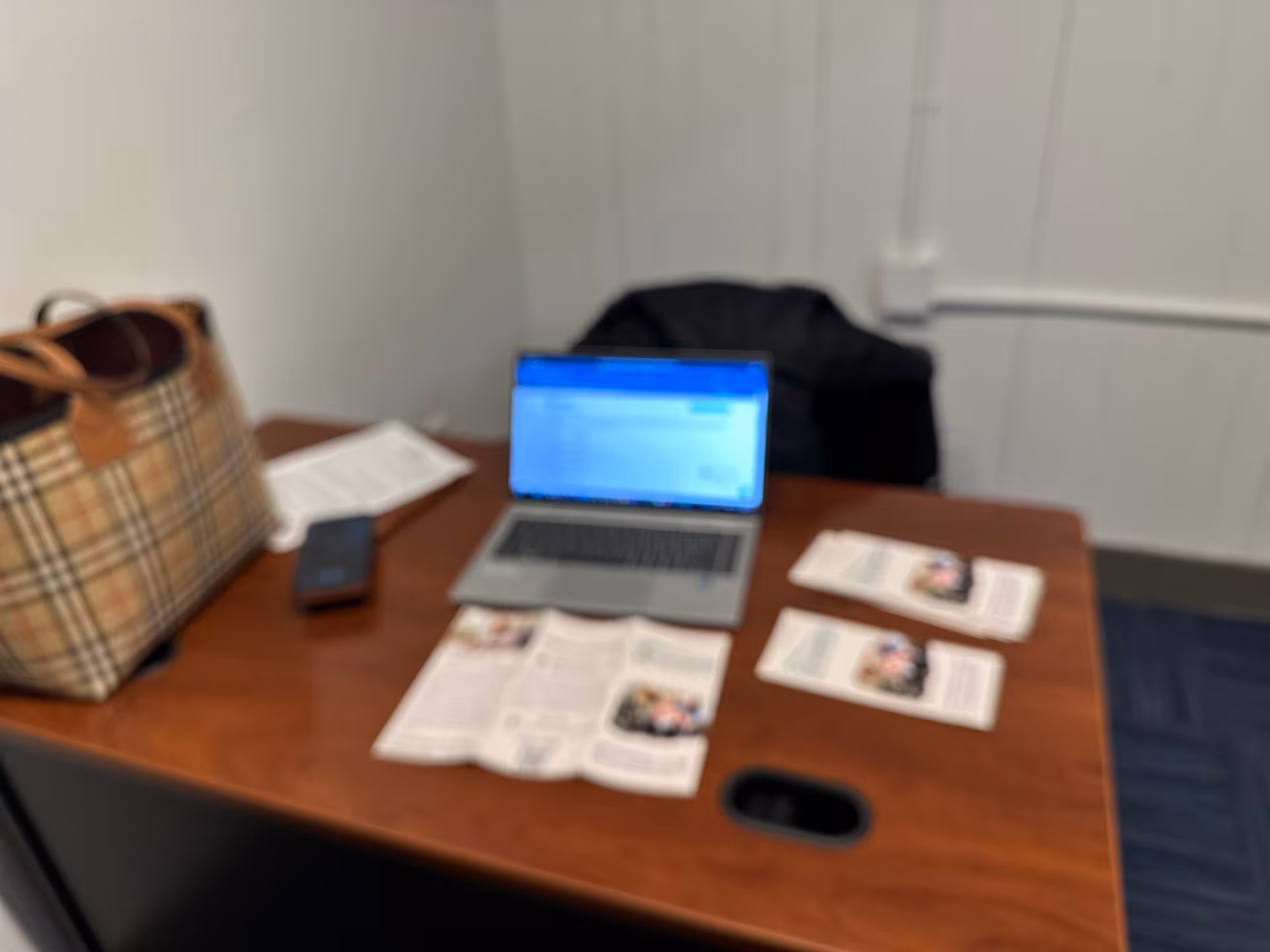 Blurry office desk with laptop, papers, and a plaid bag on a wooden table.