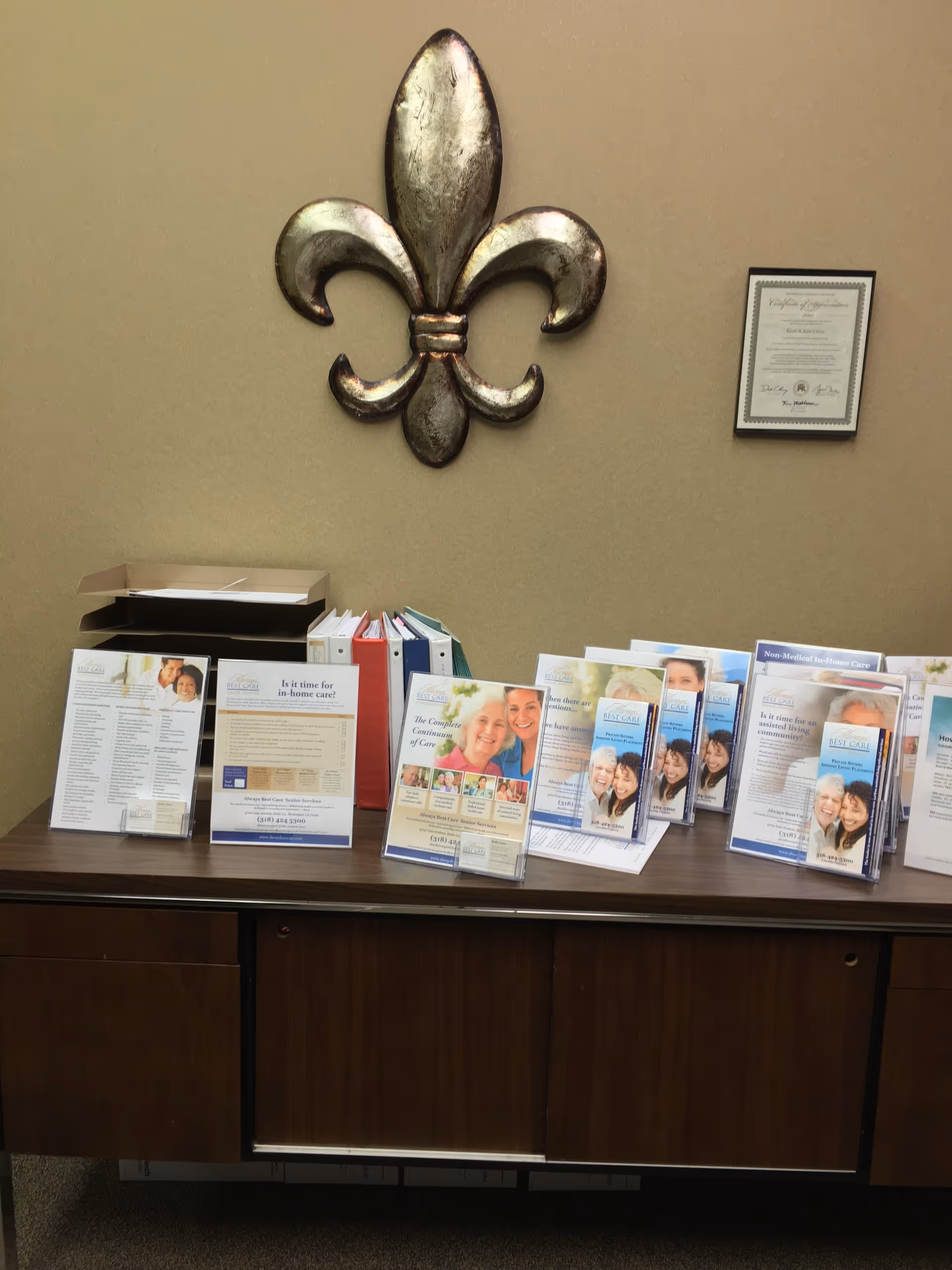 Lobby desk with brochures and fleur-de-lis wall art beside a framed certificate