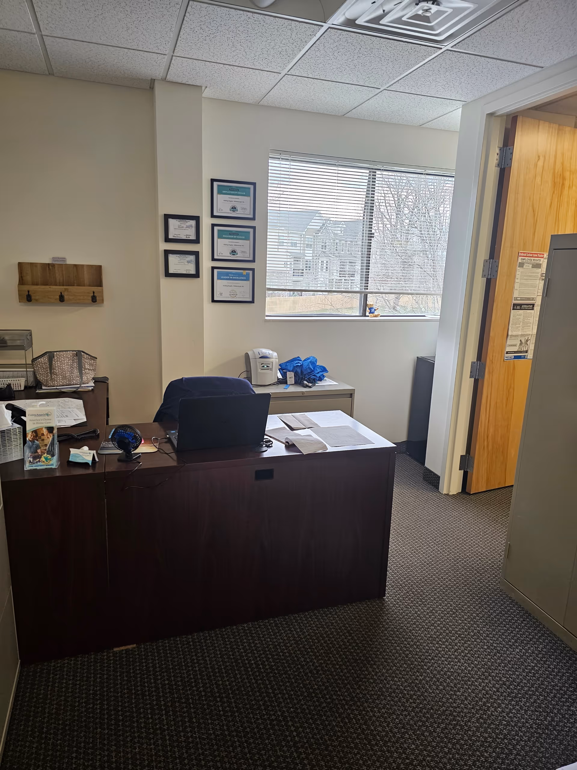 Office workspace with desk, laptop, framed certificates, and a window.