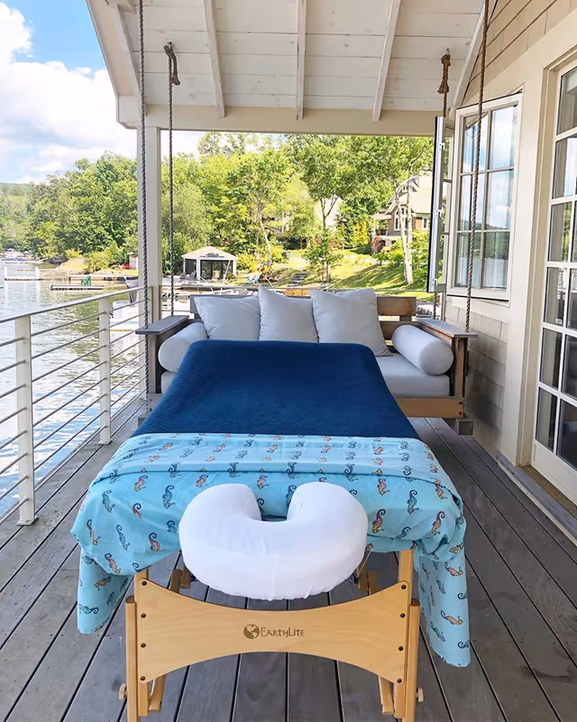 Outdoor balcony massage setup with a table, cushioned swing, and water view.