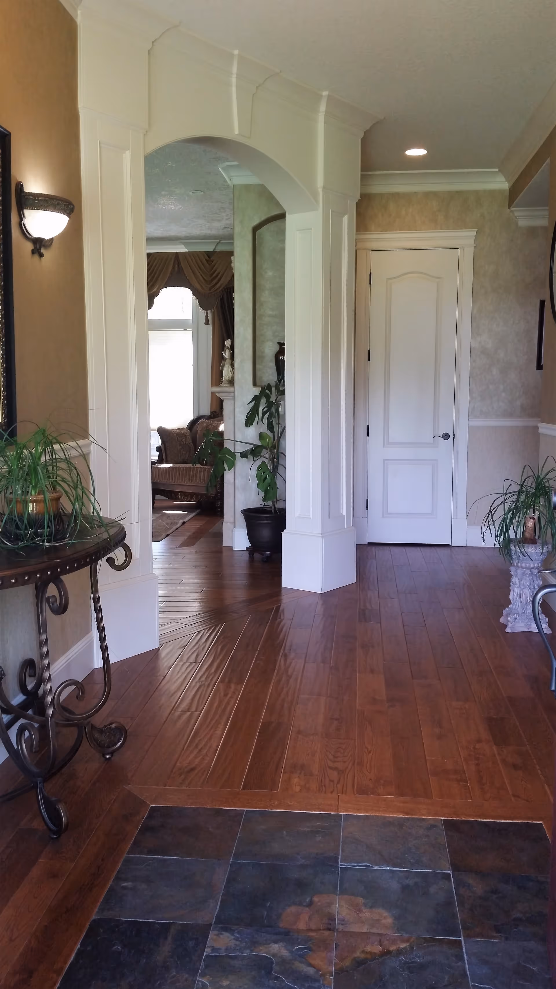 Interior foyer with archway, wooden floors, plants, and a door to another room.