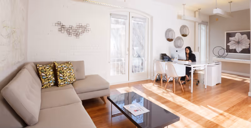 Bright living room with white walls, beige sofa, glass coffee table, and person at desk.