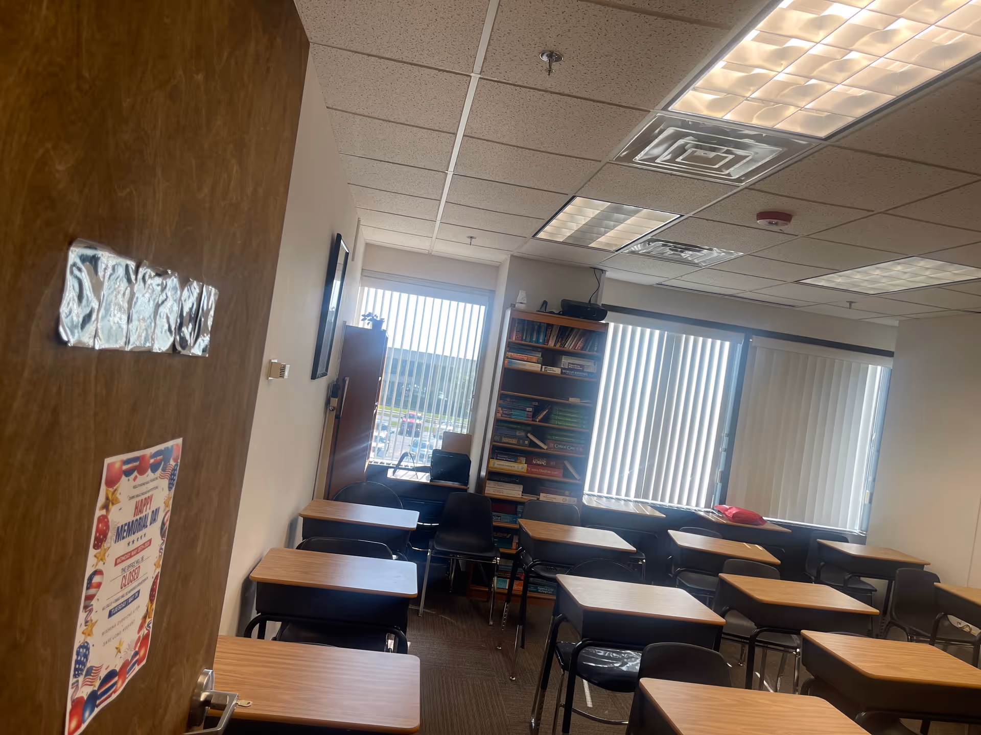Classroom-style interior with desks, chairs, a bookshelf, and large windows with blinds.