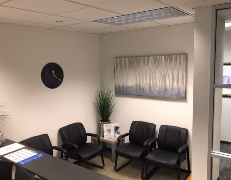 A small interior waiting area with four black chairs, plant, clock, and artwork.