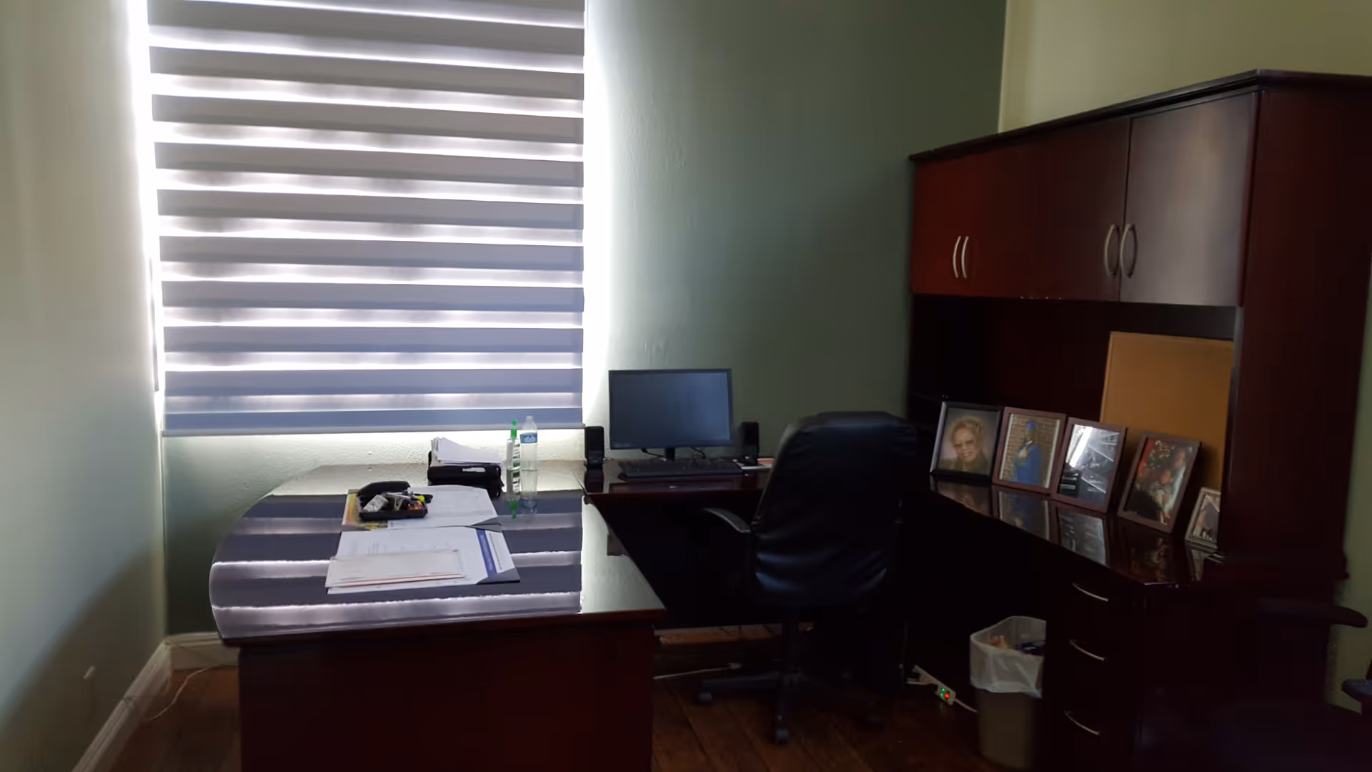 Wooden office desk with computer, papers, and framed photos in a green-walled room.
