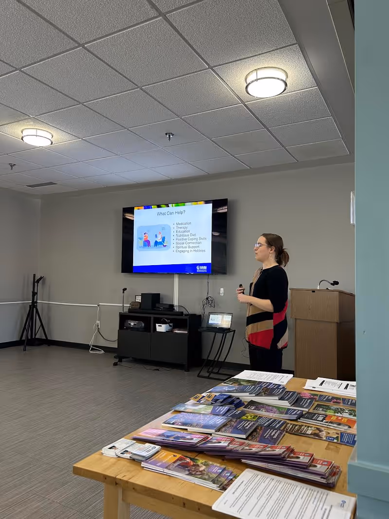 A presenter stands beside a mounted screen in a meeting room with brochures on a table.