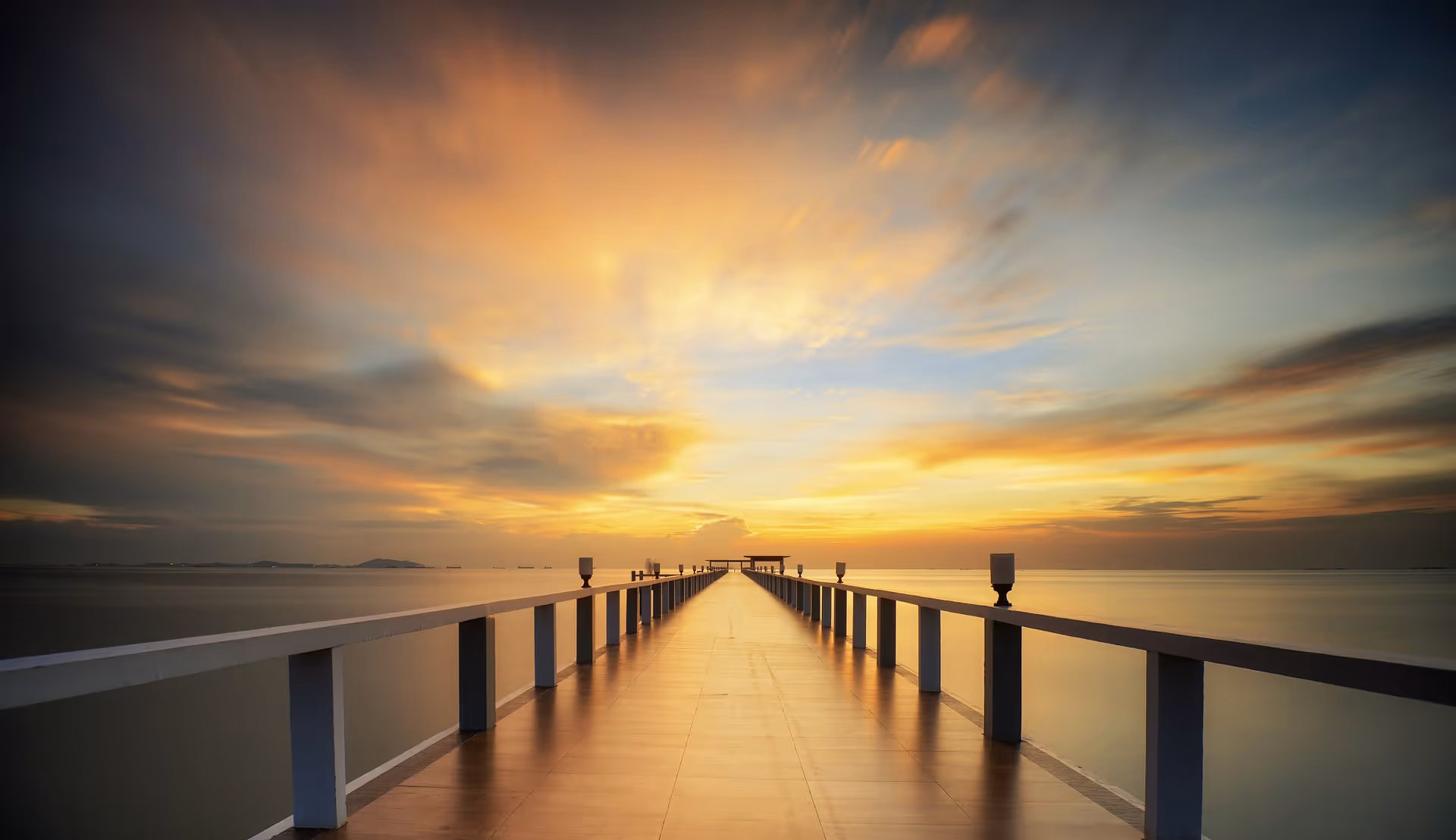 A long pier extends over calm water toward a glowing sunset.