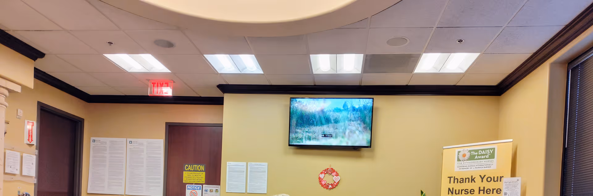 Interior waiting area with a wall-mounted TV, posters, and a nurse appreciation banner.
