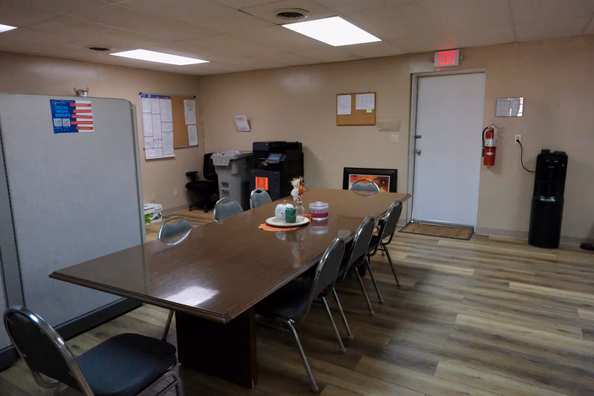 Long wooden dining table with metal chairs in a communal room; bulletin boards and copier nearby.