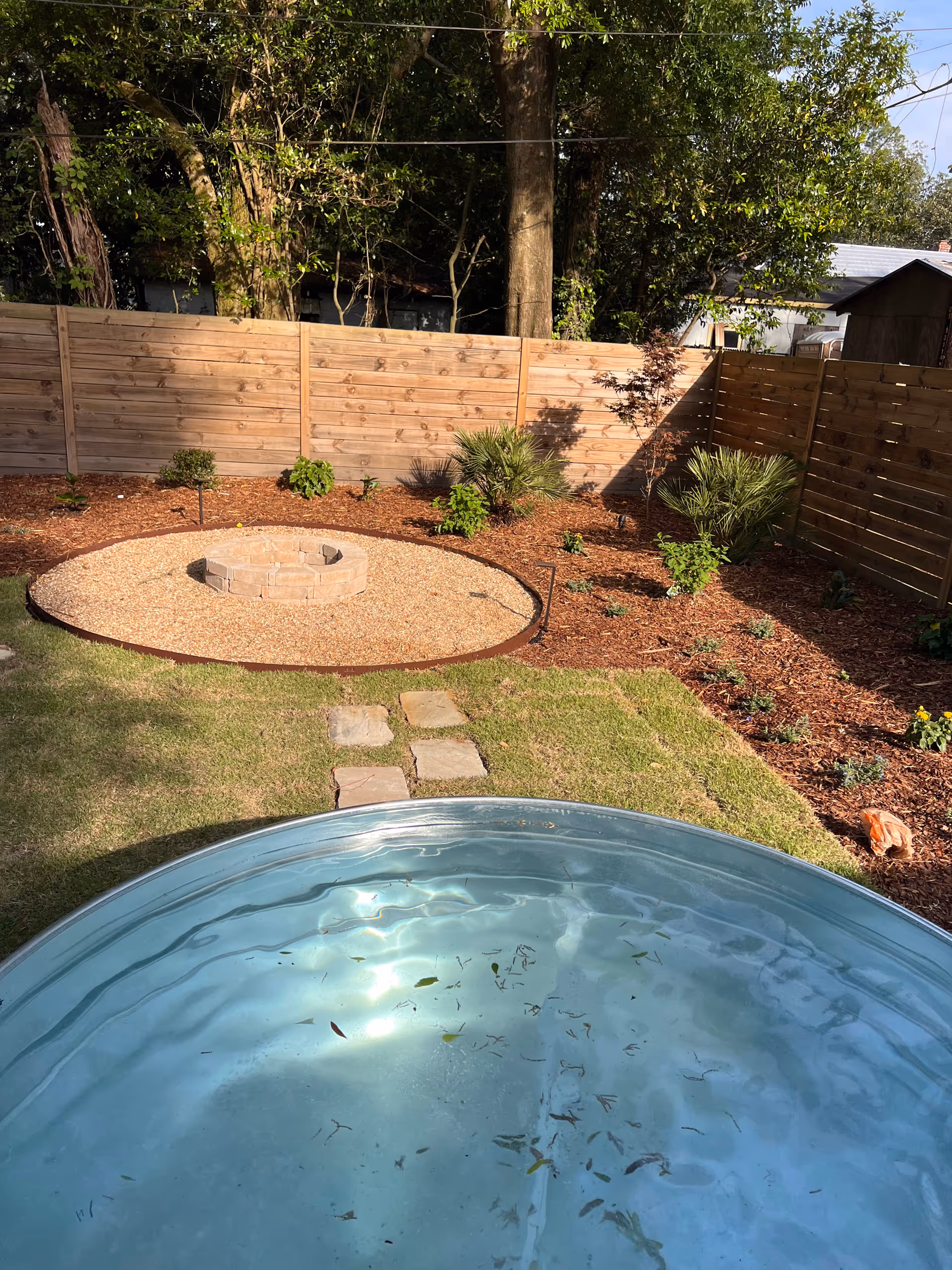 Backyard with a circular gravel fire pit, stepping stones, plants, and a round blue pool
